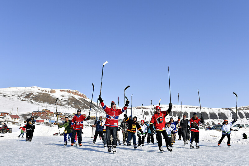 Group of ice hockey players in red and black jerseys celebrating with raised sticks on a frozen outdoor surface.
