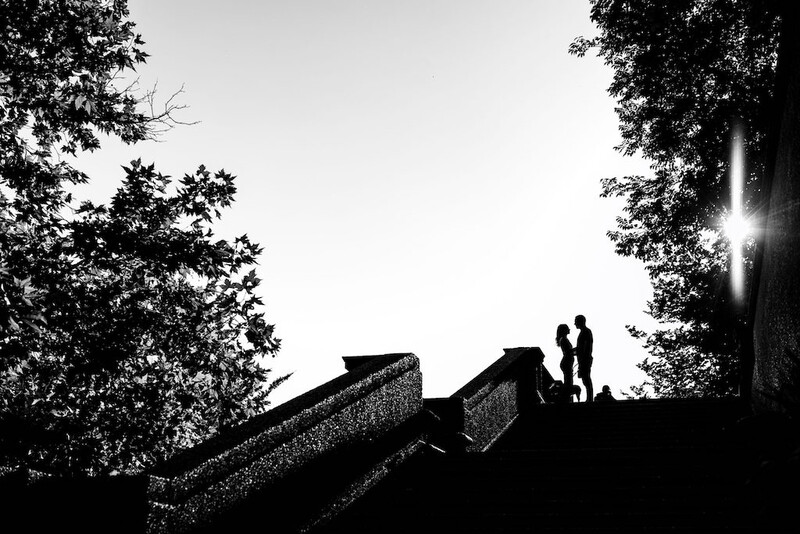 Two silhouetted figures stand atop a wide staircase against a bright sky framed by dark trees.