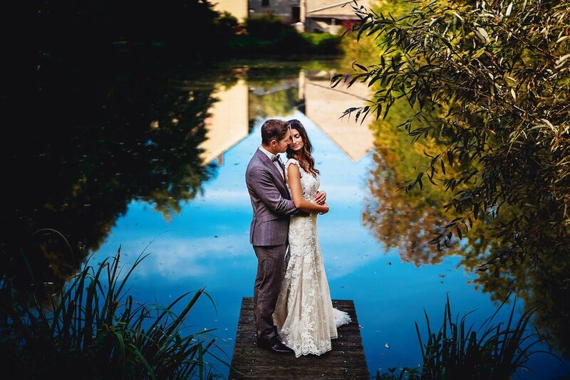 Couple embracing on a wooden dock overlooking a still blue water, framed by lush green foliage.