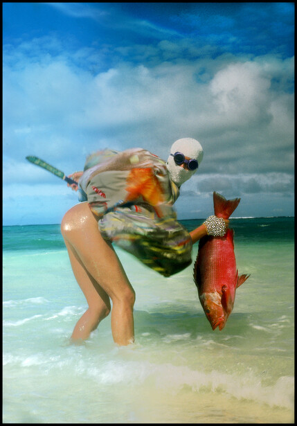 Fisherman holding large red fish in shallow ocean water with beach and blue sky behind.