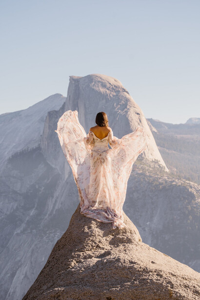 Woman in flowing dress standing on rocky outcrop with half dome mountain formation in background.
