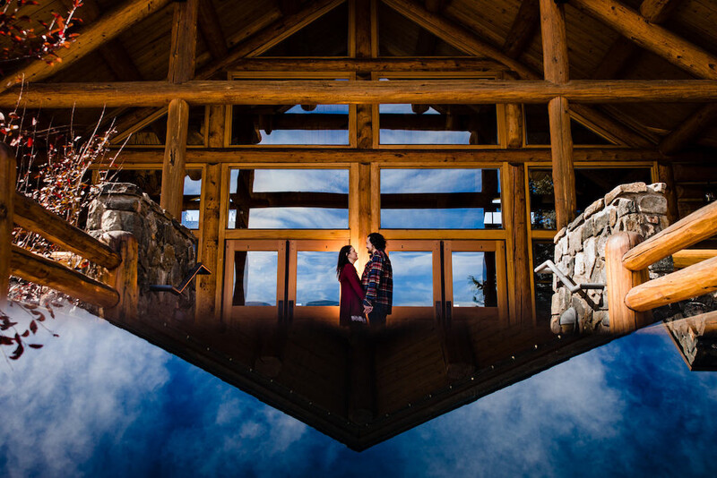 Two people silhouetted against large wooden doors and windows inside a rustic lodge, with snow-covered landscape visible through glass.