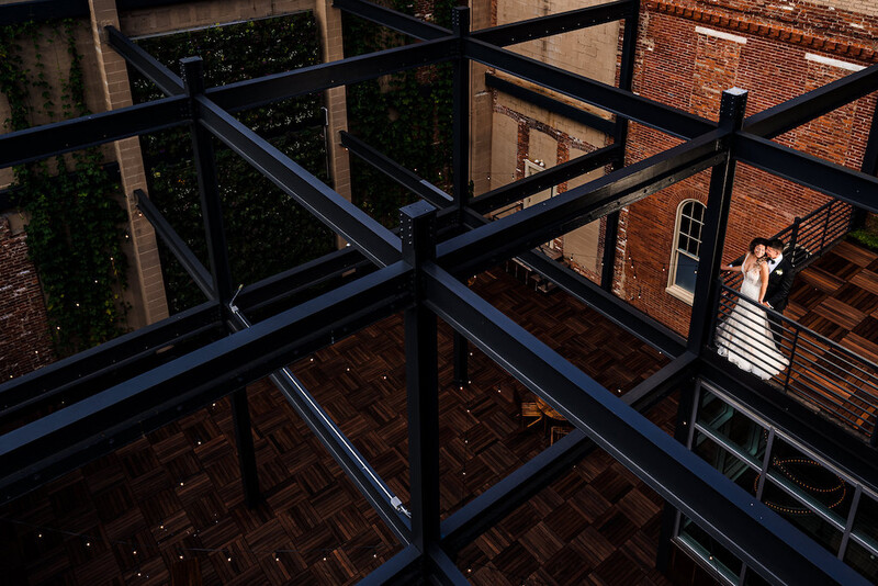 Shadow patterns from geometric metal railings cast across a brick building facade and wooden deck in harsh sunlight.