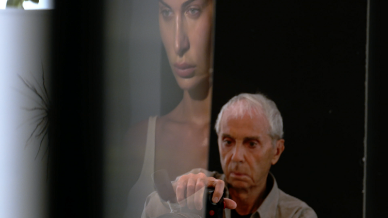 Older man with white hair speaking into a microphone, with a portrait of a woman visible in soft focus behind him.