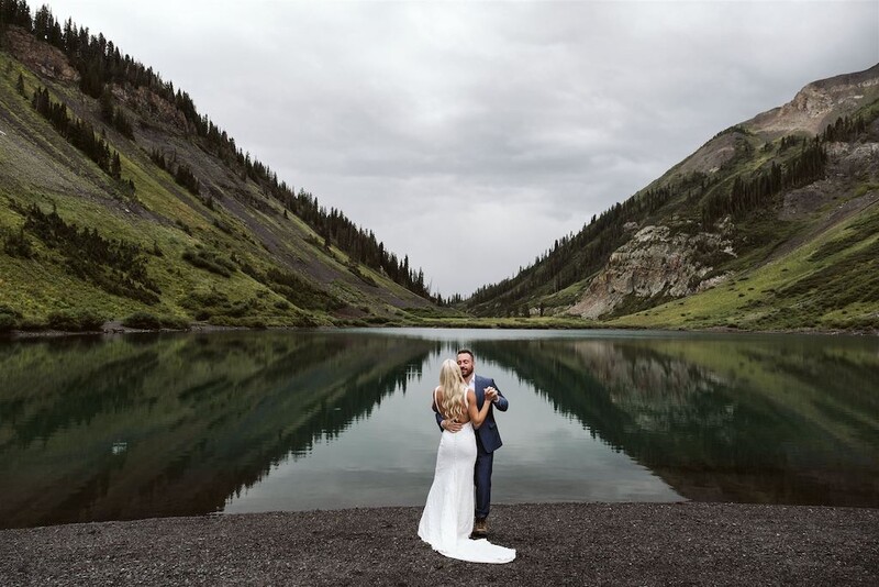 Bride and groom standing on a rocky shore facing a perfectly still lake surrounded by forested mountain slopes.