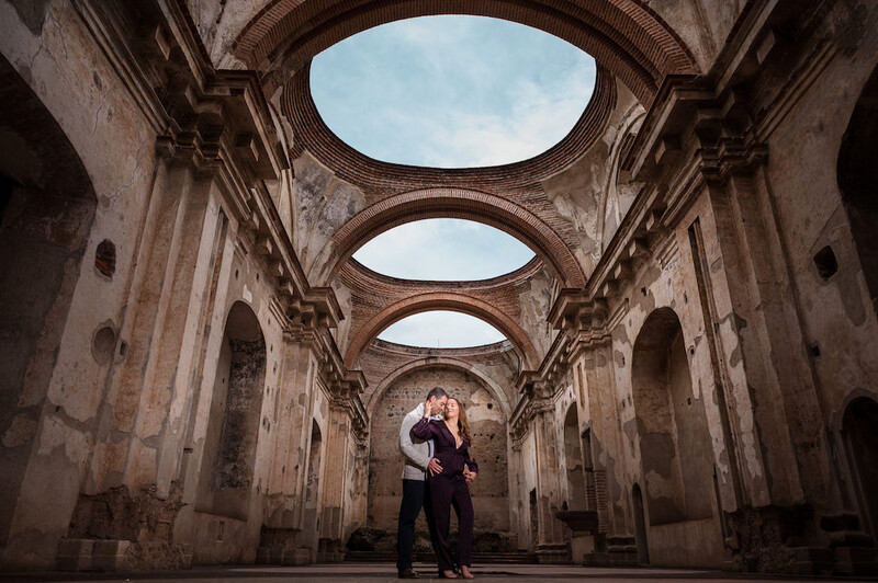 Couple embracing beneath arched ceiling with three oval skylights in an ornate, deteriorating stone corridor.