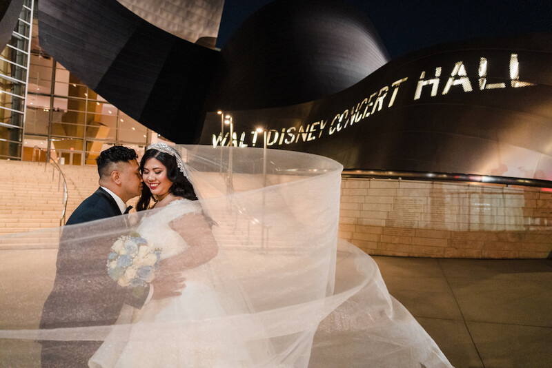 Bride and groom embrace in front of Walt Disney Concert Hall at night, with billowing veil creating movement.