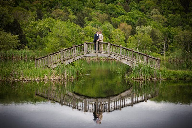Couple standing on arched wooden bridge over still water reflecting trees and sky.