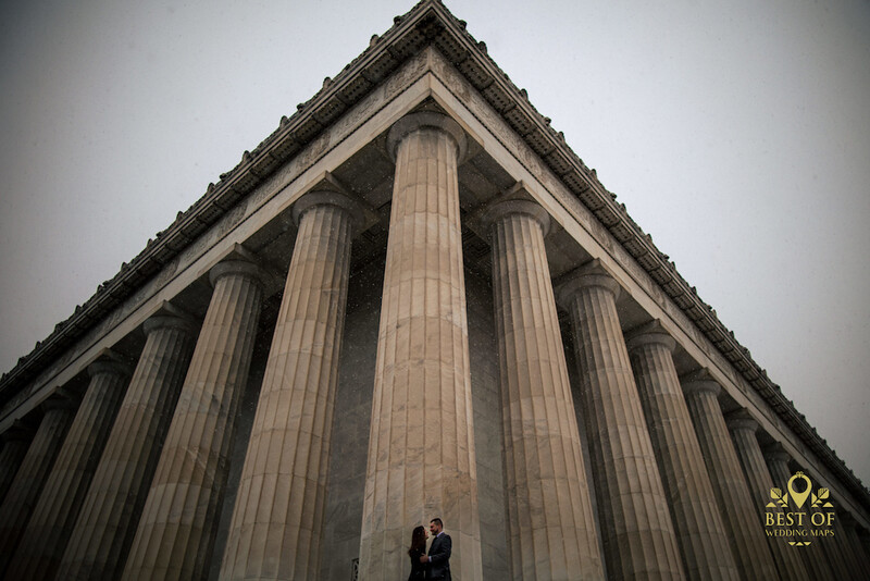 Couple standing beneath towering neoclassical columns shot from below against overcast sky.
