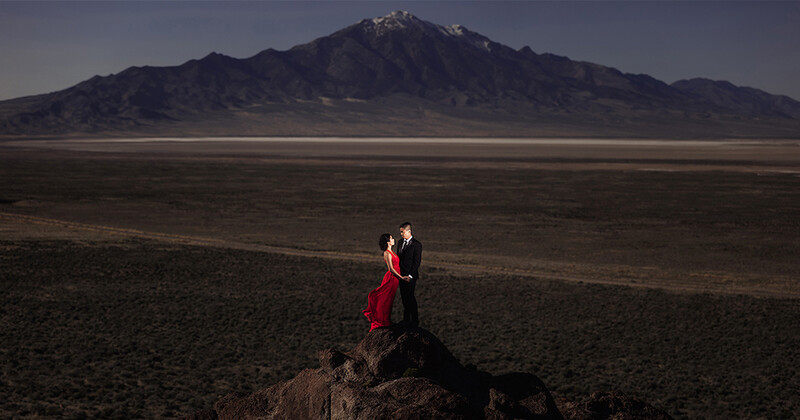 Couple in formal attire posing on a rock outcrop in an expansive desert plain with a snow-capped mountain in the distance.