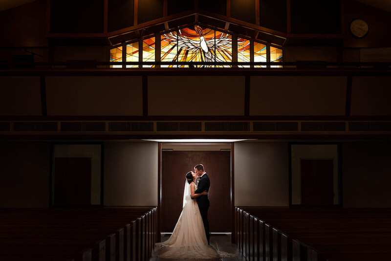 Bride and groom embrace in a doorway beneath an illuminated stained glass window in a chapel.