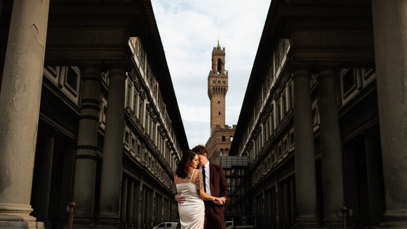 Couple kissing in a Renaissance gallery corridor with a bell tower visible in the distance.