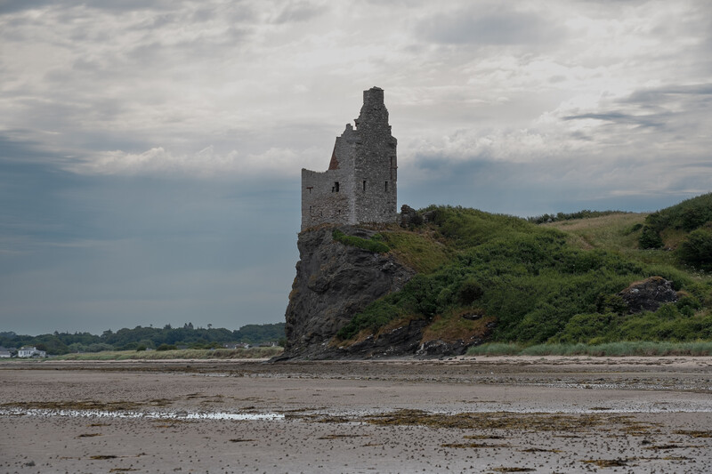 Ruined stone tower on a rocky coastal promontory at low tide under dramatic cloudy sky.