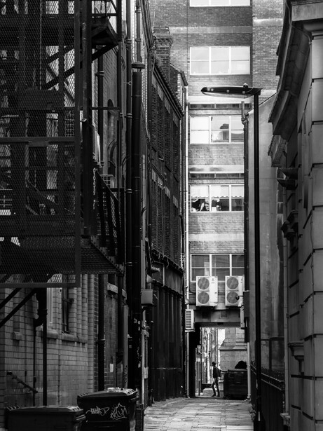 Narrow urban alley between tall buildings converging toward a distant bright window. Black and white photograph.