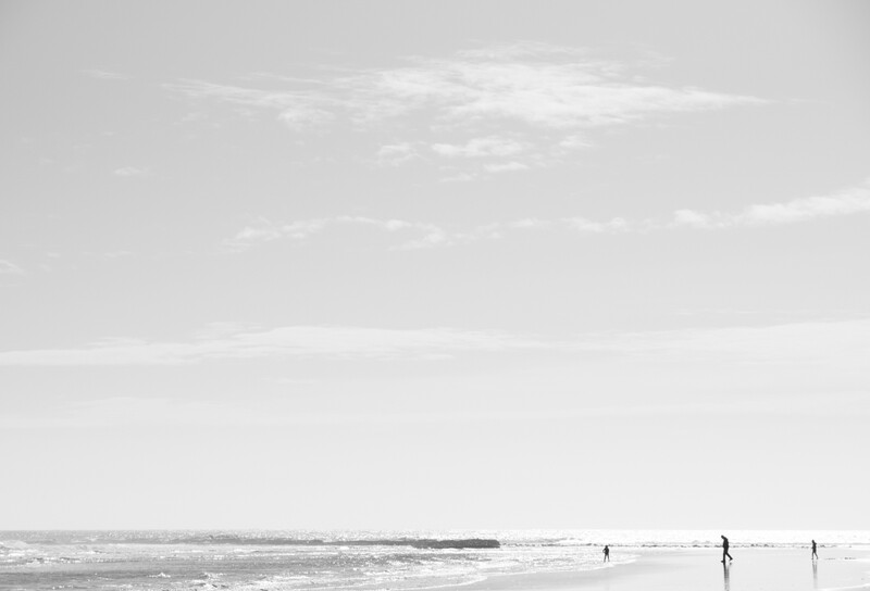 Minimalist beach scene with three solitary figures standing in shallow water under an overcast sky.