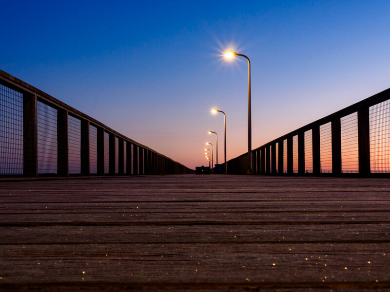Symmetrical wooden boardwalk lined with metal fencing stretches into twilight, lit by street lamps.