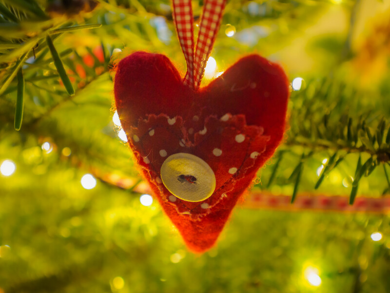 Red velvet heart ornament with polka dots and wooden button detail hanging on decorated evergreen tree with warm yellow lights.