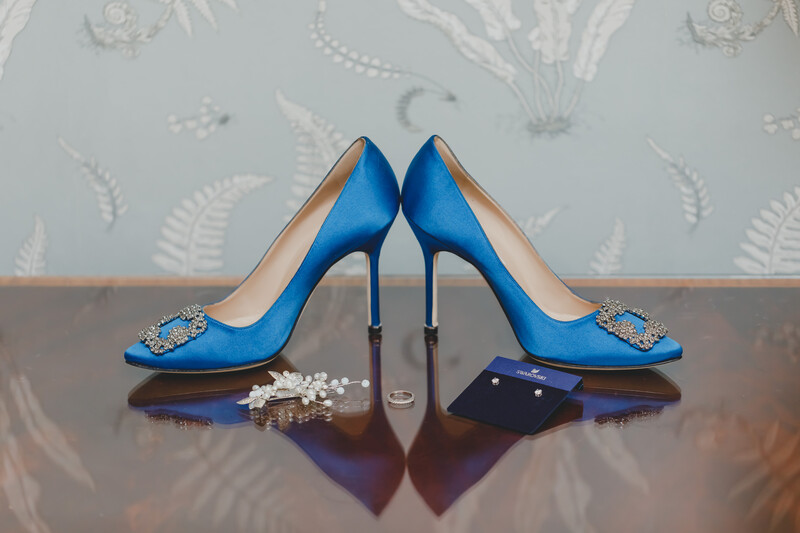Pair of bright blue satin heels with jeweled ornaments, displayed symmetrically with wedding rings and pearls on a wooden surface.