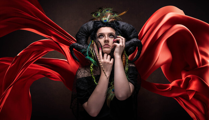 Woman with dramatic dark makeup and black hair headpiece adorned with feathers, surrounded by flowing red fabric against dark background.
