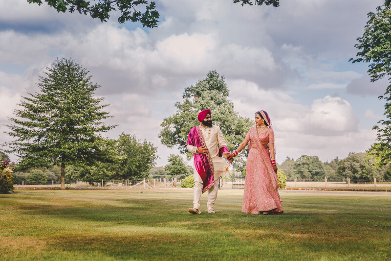 Couple in traditional South Asian wedding attire walking hand-in-hand across an open grassy field.