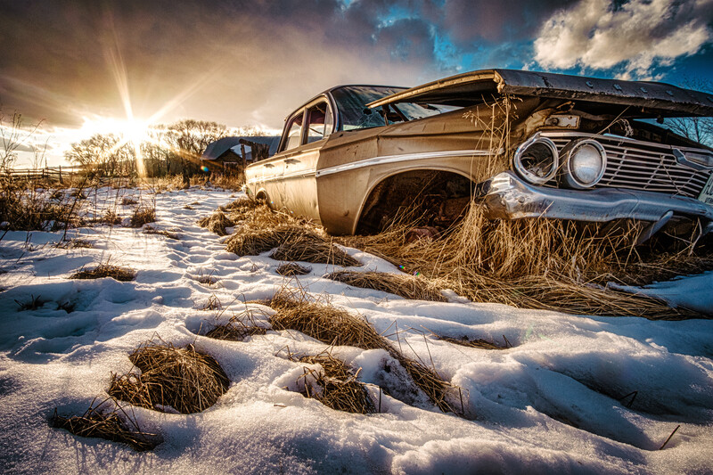 Abandoned vintage sedan half-buried in snow with dramatic golden sunlight and storm clouds overhead.