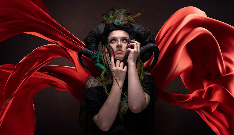Person wearing dramatic black horned headdress with green accents, surrounded by flowing red fabric against dark background.