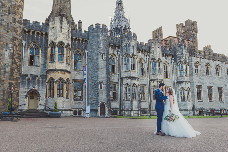 Bride and groom posing in front of a Gothic stone castle with blue-gray stonework and ornate architecture.