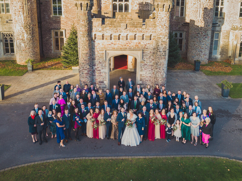 Large wedding party of approximately 80 guests posed in formation in front of a stone castle entrance.