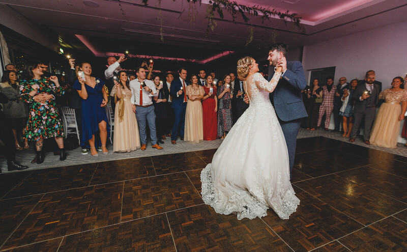 Bride and groom share their first dance on a wooden floor while wedding guests watch from the sides of the venue.