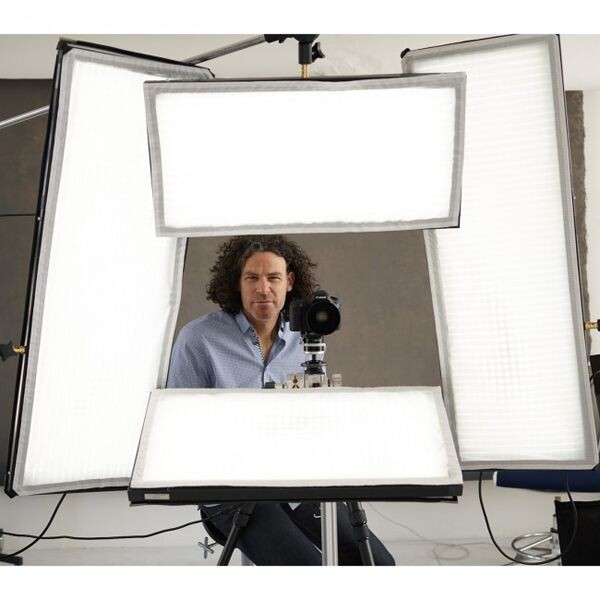 Man with curly hair positioned behind a large square softbox lighting setup with camera on tripod.