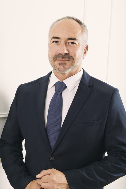 Professional headshot of a man in a dark blazer and blue tie against a neutral background.