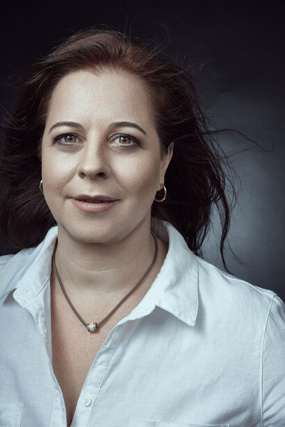 Woman with dark hair wearing a light blue collared shirt against a dark background.