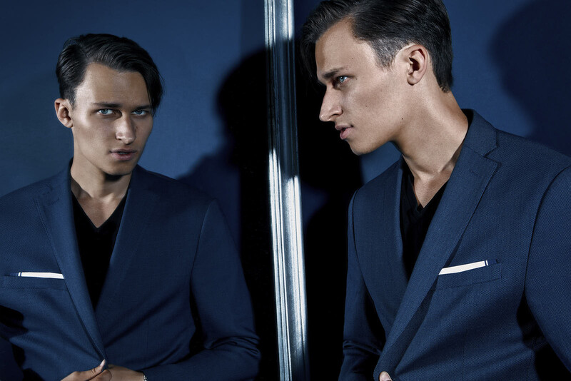 Two men in dark blazers facing each other across a reflective vertical surface against a blue background.