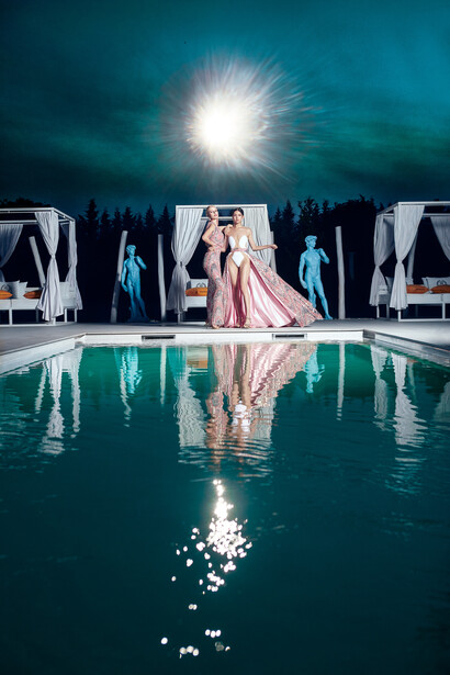 Woman in flowing pink gown reflected in still pool water at night under bright moon.