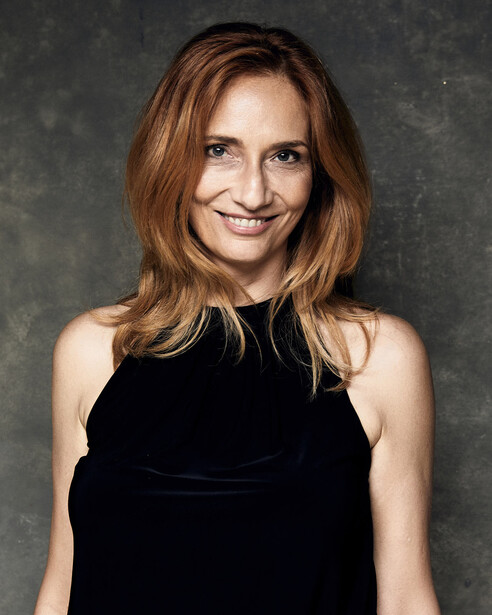 Woman with shoulder-length auburn hair smiling at camera against neutral gray background.