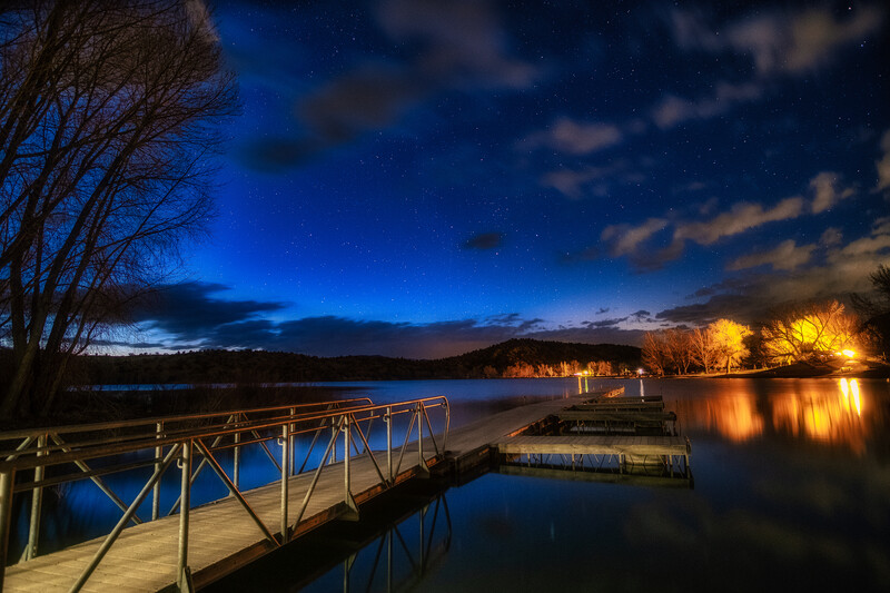 Wooden dock extending into a calm lake at twilight, with warm lights reflecting on water and trees framing the scene.