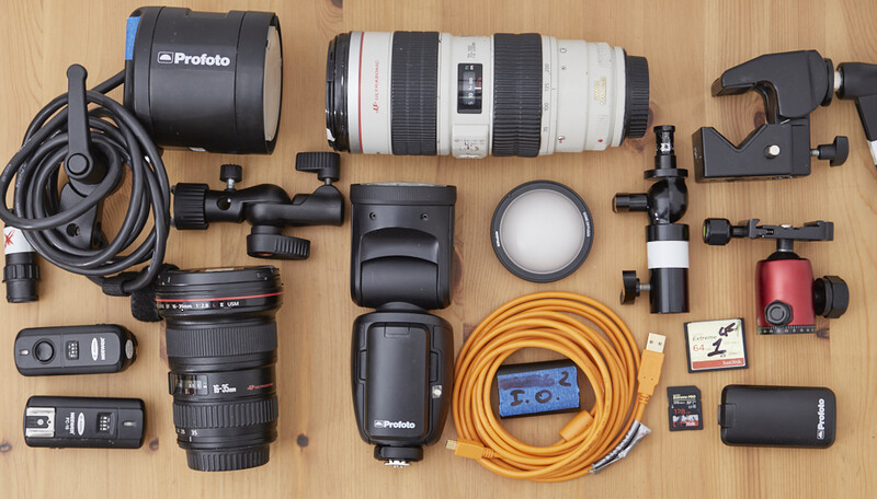 Professional photography gear laid out flat on wooden surface, including lenses, lighting equipment, and wireless triggers.