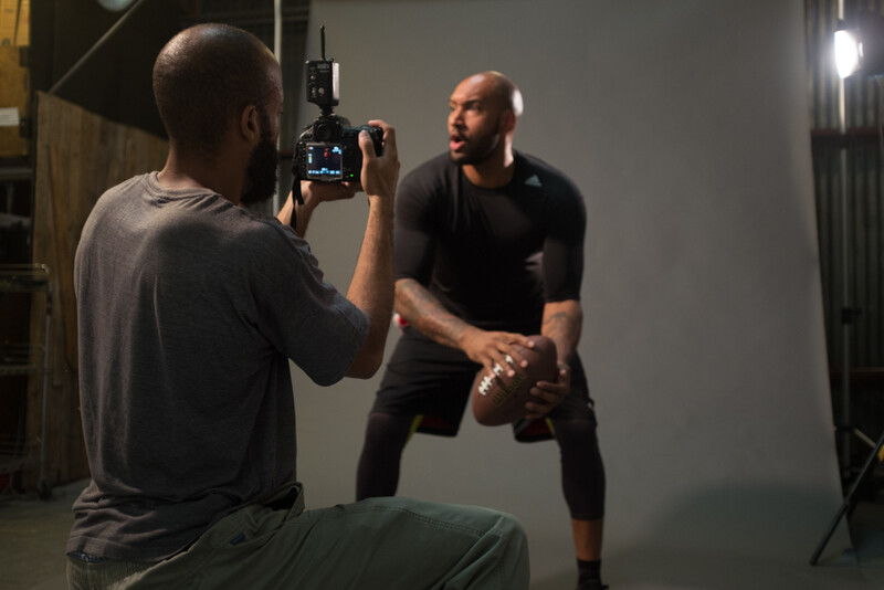Photographer operating a cinema camera on a tripod while recording a male subject in a studio setting with professional lighting.