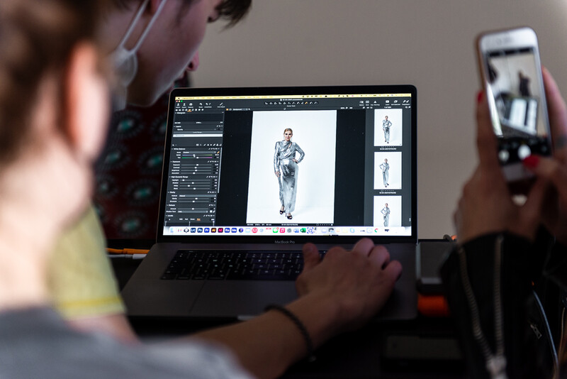 Photographer editing fashion photos on laptop while using smartphone to reference poses.