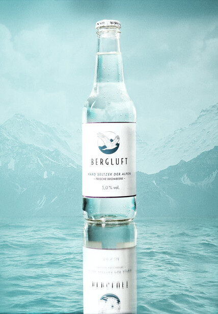 Product photography of a clear glass water bottle with white label, reflected in still water against a misty mountain backdrop.