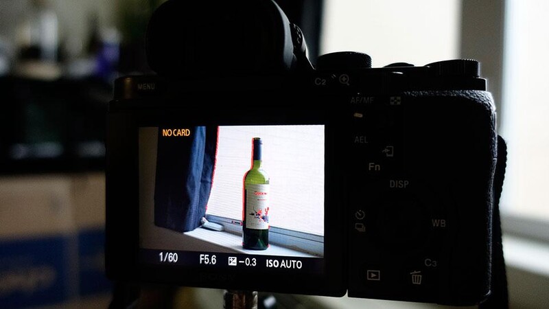 Camera LCD screen displaying exposure settings while framing a wine bottle against a window.