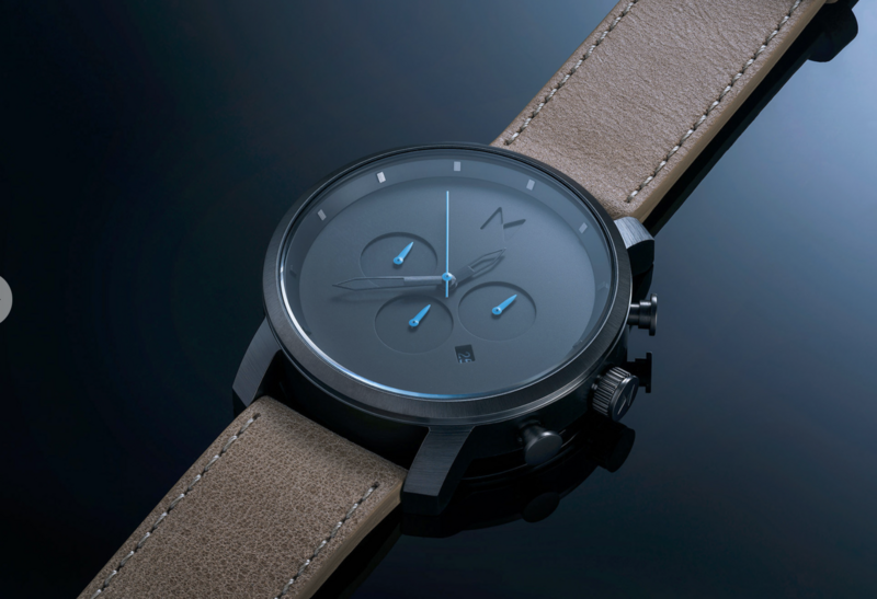 Modern chronograph watch with dark blue dial, blue subdial accents, and tan leather strap against dark background.