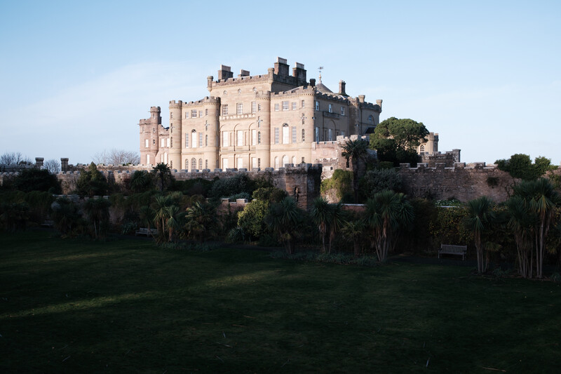 Medieval castle with stone towers and crenellations perched on a hilltop, surrounded by green grounds and palm trees.