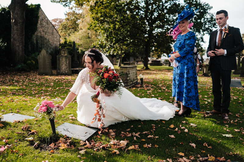 Bride in white dress kneeling in cemetery holding bouquet while guests in formal attire stand nearby.