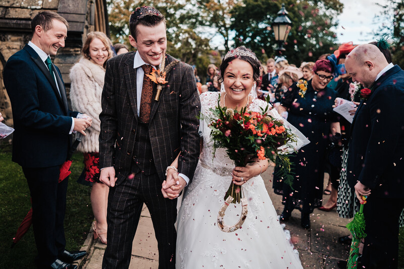 Newlyweds walk together down an outdoor path while confetti falls around them.