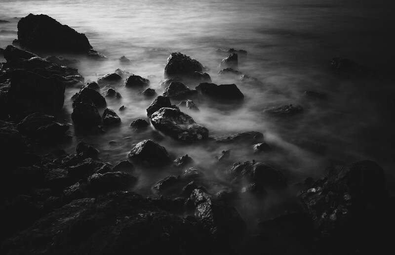 Rocky coastline shrouded in misty seawater with long exposure technique creating ghostly waves around dark boulders.