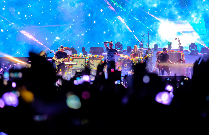 Concert stage with performers silhouetted against vibrant blue and purple lighting, audience in foreground with raised phones creating bokeh light spots.