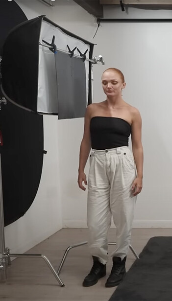 Model standing in studio with professional lighting setup and reflector visible in background.