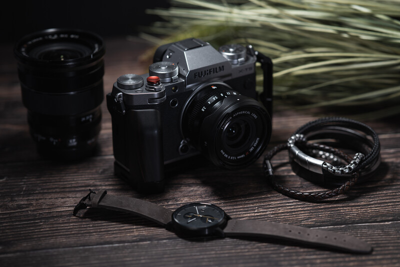 Vintage film camera with interchangeable lenses, watch, and jewelry arranged on dark wood surface.