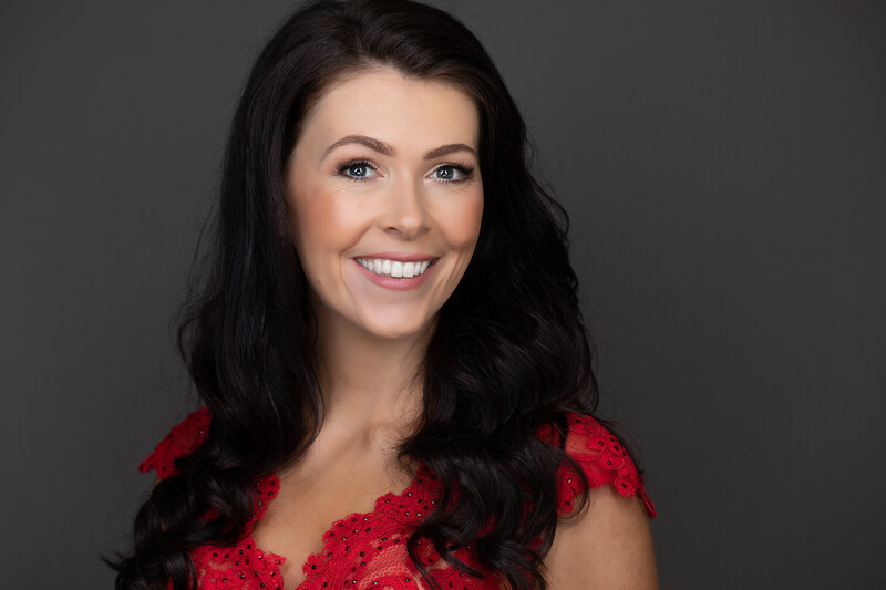 Woman with long black hair wearing a red lace top, smiling at camera against neutral gray background.